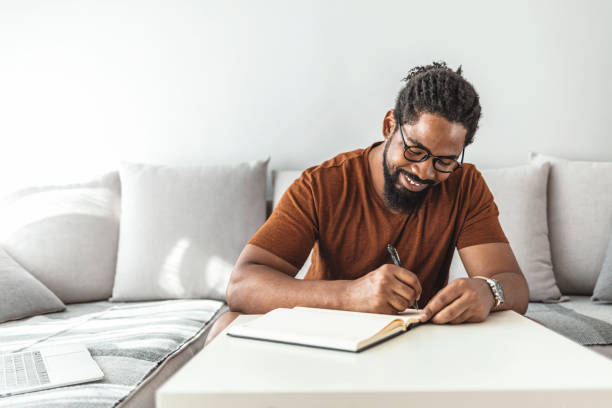 Image Of Happy African Man With Notepad And Pen Sitting On Sofa.