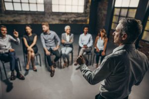 Mid Adult Instructor Talking To Large Group Of Business People On A Therapy.