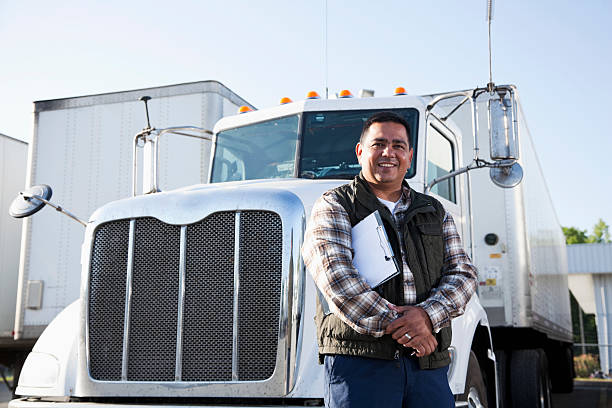 Hispanic Truck Driver (40s) Standing In Front Of Semi Truck With Clipboard.